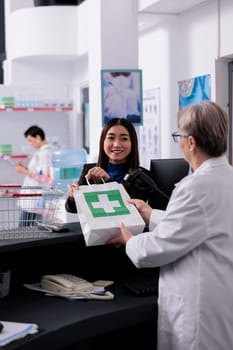 Woman chatting with pharmacy cashier at chekout, taking shopping bag with medical products. Elderly drugstore worker giving young asian customer medicaments online order package at counter desk