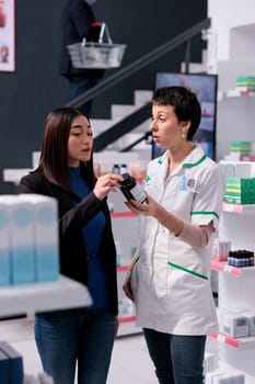 Young woman discussing multivitamin choice with drugstore assistant. Pharmaceutical employee wearing medical worker uniform advising asian client nutritional element pills in apothecary