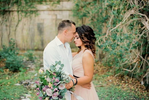Groom hugs bride touching her forehead with his forehead near the ancient stone wall. High quality photo