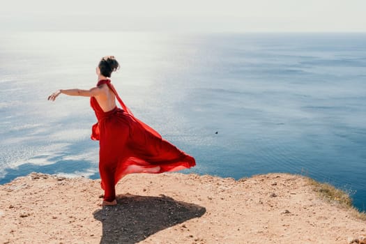 Side view a Young beautiful sensual woman in a red long dress posing on a rock high above the sea during sunrise. Girl on the nature on blue sky background. Fashion photo.