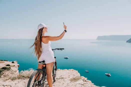 A woman cyclist on a mountain bike looking at the landscape of mountains and sea. Rear view of cyclist woman standing in front to the sea with outstretched arms. Freedom and healthy lifestyle concept