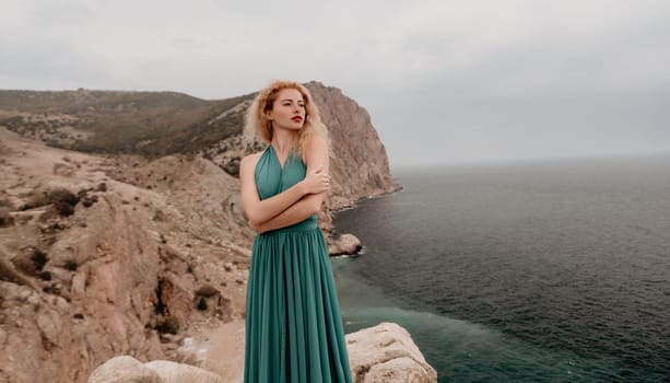 Side view a Young beautiful sensual woman in a mint long dress posing on a volcanic rock high above the sea during sunset. Girl on the nature on overcast sky background. Fashion photo