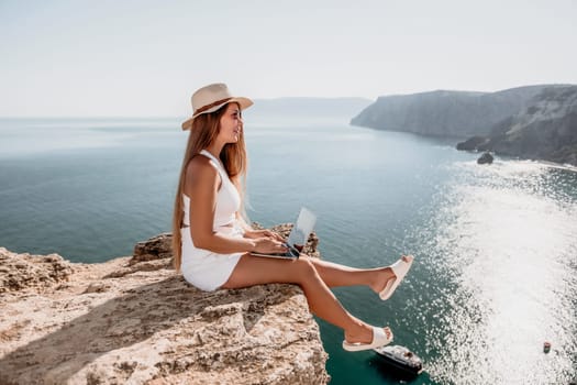 Successful business woman in yellow hat working on laptop by the sea. Pretty lady typing on computer at summer day outdoors. Freelance, travel and holidays concept.