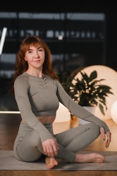 A woman in a gray tracksuit sits on a yoga mat in a lotus position in the gym.