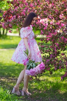 a girl with long hair, in a pink dress, standing with a large bouquet of peonies on green grass, in a blooming garden. Vertical. Copy space