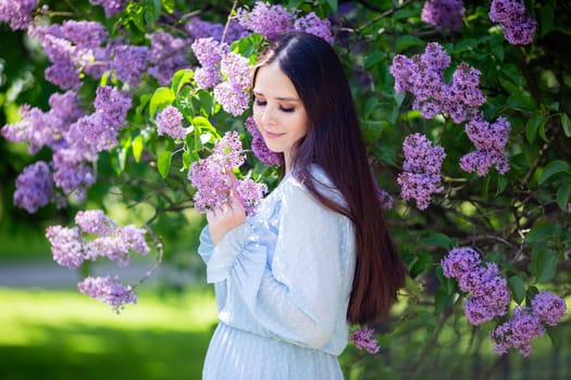 A cute enjoying girl in light blue dress, stands next to a flowering lilac bush in the park. Looking down. Close up. Copy space