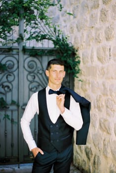 Groom in a vest with a jacket on his shoulder stands in the courtyard of an old stone building. High quality photo