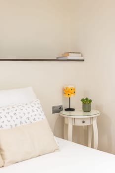 Close-up corner of cozy bedroom in light white colors with bedside table with lamp and shelf next to the bed with linen and a pillow. Concept of a calm and concise interior.