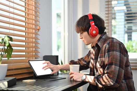 Hipster young asian man wearing wireless headphone and browsing internet in digital tablet.