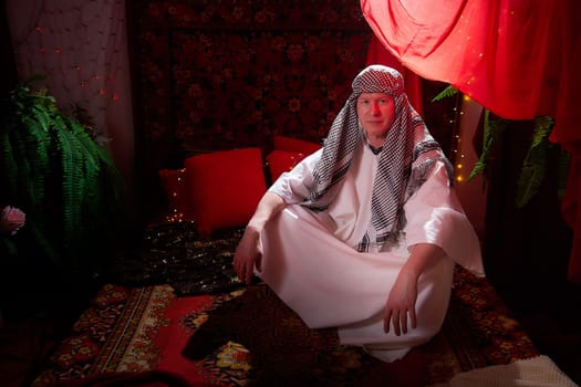 Arab sheikh in a white robe and a headscarf in a boudoir in a room with a red carpet. Oriental eastern fabulous style photo shoot with man. Male model posing in fary tale