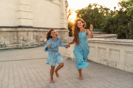 Mother's daughter sunset. in blue dresses with loose long hair against the background of a sunset and a white building. They run and hold hands