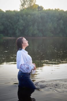 Portrait of beautiful young woman in stylish dress with white shirt and jeans in water of river or lake in nature landscape in summer evening