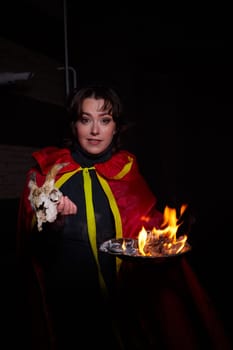 Witch woman in red cloak with skull on black background and light in night. Female warrior in Dark