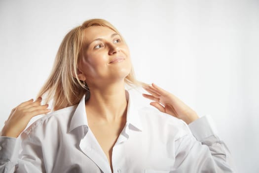 Portrait of a pretty blonde smiling woman posing on white background and correcting her hair and hairstyle. Happy girl model in white shirt in studio