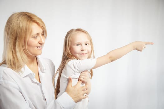 Portrait of a blonde mother and daughter who having communicate and play on white background. Mom and little girl models pointing his finger forward. The concept of family unity
