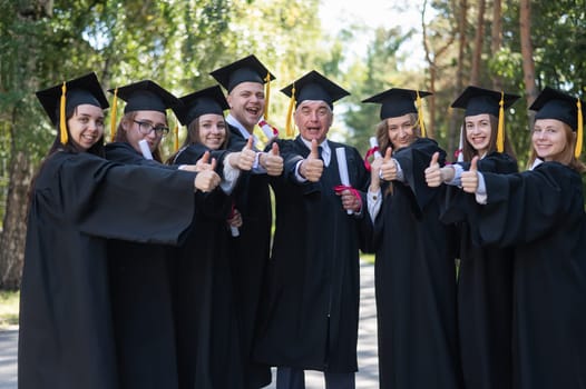 A group of graduates in robes give a thumbs up outdoors. Elderly student