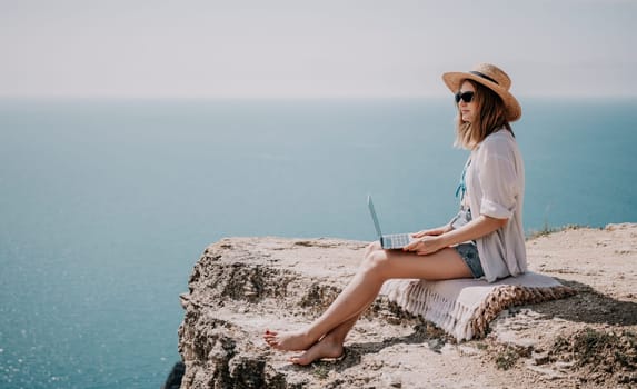 Successful business woman in yellow hat working on laptop by the sea. Pretty lady typing on computer at summer day outdoors. Freelance, travel and holidays concept.