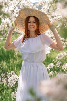 Woman cherry orchard. A happy woman in a long white long dress walks through the green spring blooming cherry garden. Happy cheerful princess bride. The fabric of the skirt flutters in the wind