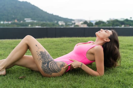 A slender young woman in a bright swimsuit on the grass by the pool, a large tattoo on her thigh. Slim athletic girl posing, tender romantic sexy