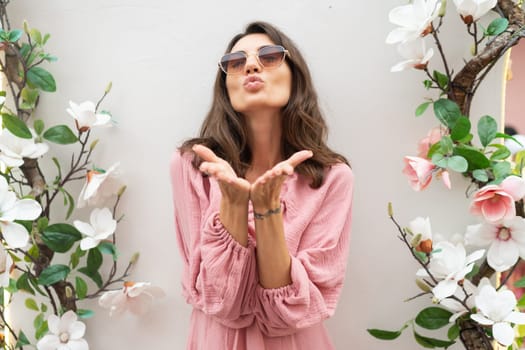 Young beautiful smiling cute romantic woman in trendy summer pink dress. Carefree woman posing in the street near white wall with flowers. Positive model outdoors in sunglasses.