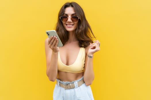 Young beautiful smiling woman with mobile phone. Carefree woman posing in the street near yellow wall. Positive model outdoors in sunglasses. Happy and excited