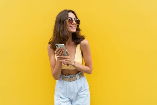 Young beautiful smiling woman with mobile phone. Carefree woman posing in the street near yellow wall. Positive model outdoors in sunglasses. Happy and excited