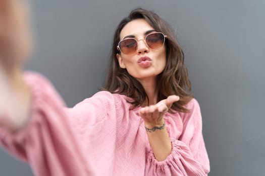 Young beautiful smiling cute romantic woman in trendy summer dress. Carefree woman posing in the street near grey wall. Positive model outdoors in sunglasses. Takes selfie.