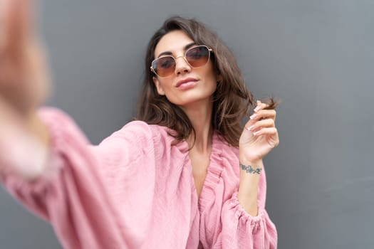 Young beautiful smiling cute romantic woman in trendy summer dress. Carefree woman posing in the street near grey wall. Positive model outdoors in sunglasses. Takes selfie.