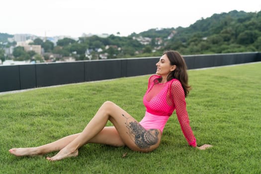 A slender young woman in a bright swimsuit on the grass by the pool, a large tattoo on her thigh. Slim athletic girl posing, tender romantic sexy