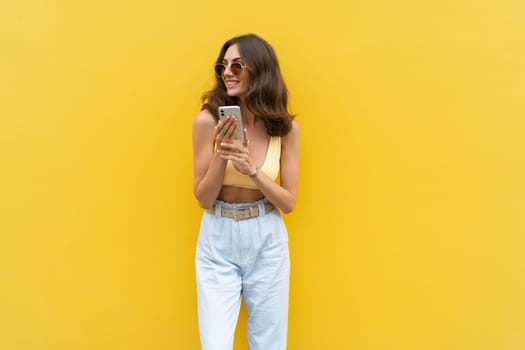 Young beautiful smiling woman with mobile phone. Carefree woman posing in the street near yellow wall. Positive model outdoors in sunglasses. Happy and excited