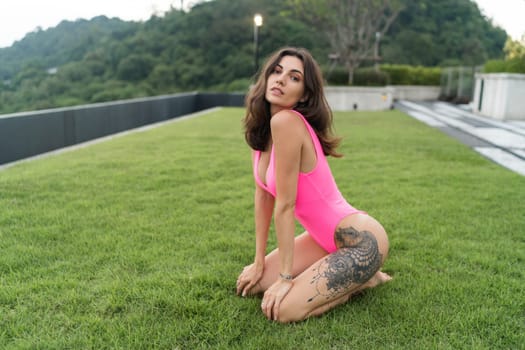 A slender young woman in a bright swimsuit on the grass by the pool, a large tattoo on her thigh. Slim athletic girl posing, tender romantic sexy