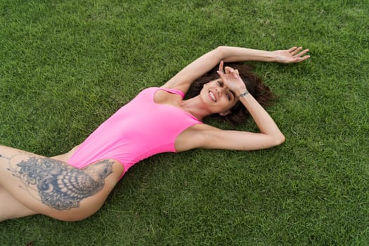 A slender young woman in a bright swimsuit on the grass by the pool, a large tattoo on her thigh. Slim athletic girl posing, tender romantic sexy