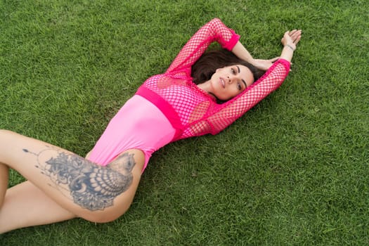 A slender young woman in a bright swimsuit on the grass by the pool, a large tattoo on her thigh. Slim athletic girl posing, tender romantic sexy