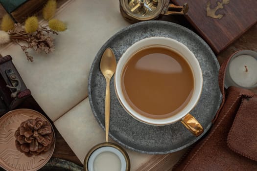 Hot coffee in ceramic cup and brass coffee spoon served with Fresh milk on worktable with old book, brass compass, scented candle and dried flowers. Space for text, Top view, Selective focus.