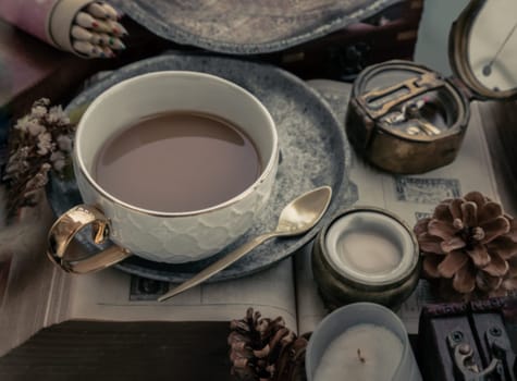 Hot coffee in ceramic cup and brass coffee spoon served with Fresh milk on worktable with old book, brass compass, scented candle and dried flowers. Selective focus.