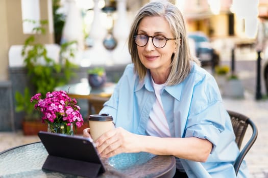 Silver-haired mature European woman sitting at an outdoor cafe, smiling with joy as she looks at her tablet PC. The image captures the happiness and contentment of a retiree enjoying leisure time and staying connected with the world through technology. High quality photo