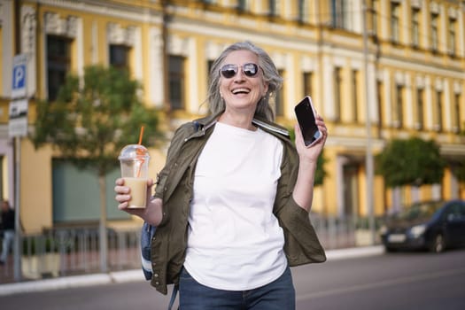 A Mature European Woman Enjoys Life Walking in The City. Biohacking Concept. Mature gray-haired modern european woman walks around the city in sunglasses, holding a mobile phone and a drink with a straw. High quality photo.