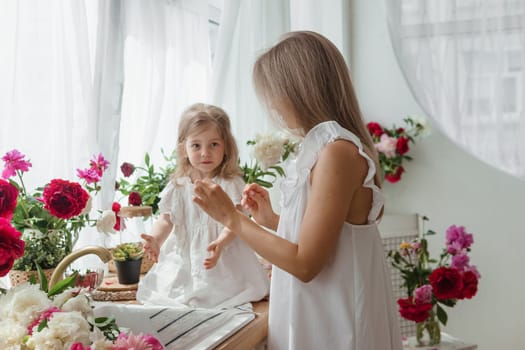 A little blonde girl with her mom on a kitchen countertop decorated with peonies. The concept of the relationship between mother and daughter. Spring atmosphere.