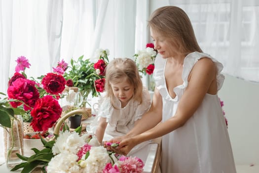 A little blonde girl with her mom on a kitchen countertop decorated with peonies. The concept of the relationship between mother and daughter. Spring atmosphere.