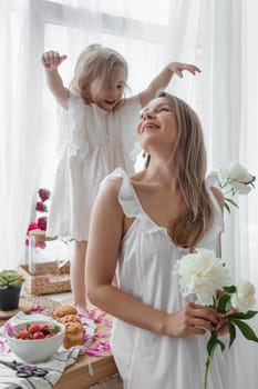 A little blonde girl with her mom on a kitchen countertop decorated with peonies. The concept of the relationship between mother and daughter. Spring atmosphere.