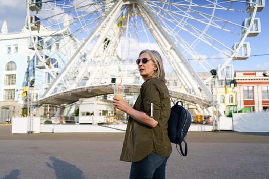 Stylish and confident mature woman with grey hair enjoys refreshing juice cup in vibrant European city, exuding elegance and positivity. The image captures the joy and active lifestyle of mature woman who is still fashion-savvy and full of life. High quality photo