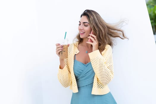 Portrait of beautiful woman in green summer dress on white background natural daylight, holding iced coffee cappuccino talking on phone smiling