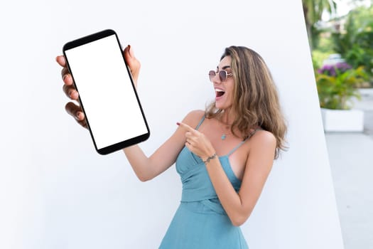 Portrait of beautiful woman in green summer dress on white background natural daylight, pointing on empty white big screen of smartphone, space for advertising
