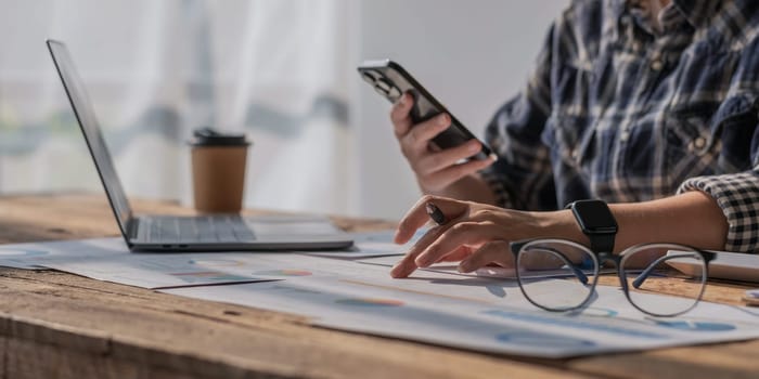 Young accountant hand holding smartphone to call marketing consultants and using laptop computer to analyze sales growth in the global workplace market. accounting concept..