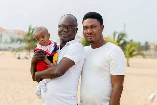a young man in glasses and dressed in white shirt standing with his friend at the beach holding his baby sticking to his chest and look at the camera.