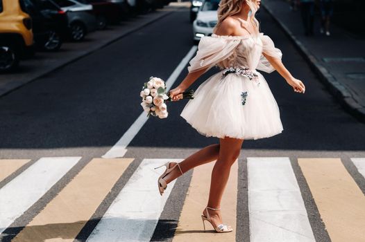 an unrecognizable bride in a short white wedding dress with a bouquet runs across the road in the city at a pedestrian crossing.