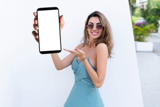 Portrait of beautiful woman in green summer dress on white background natural daylight, pointing on empty white big screen of smartphone, space for advertising