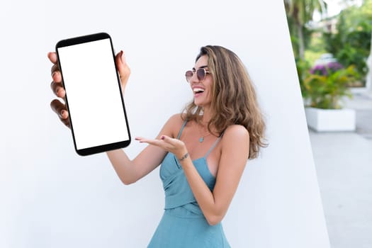 Portrait of beautiful woman in green summer dress on white background natural daylight, pointing on empty white big screen of smartphone, space for advertising