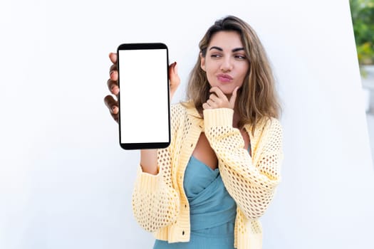 Portrait of beautiful woman in green summer dress on white background natural daylight, pointing on empty white big screen of smartphone, space for advertising