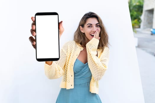 Portrait of beautiful woman in green summer dress on white background natural daylight, pointing on empty white big screen of smartphone, space for advertising